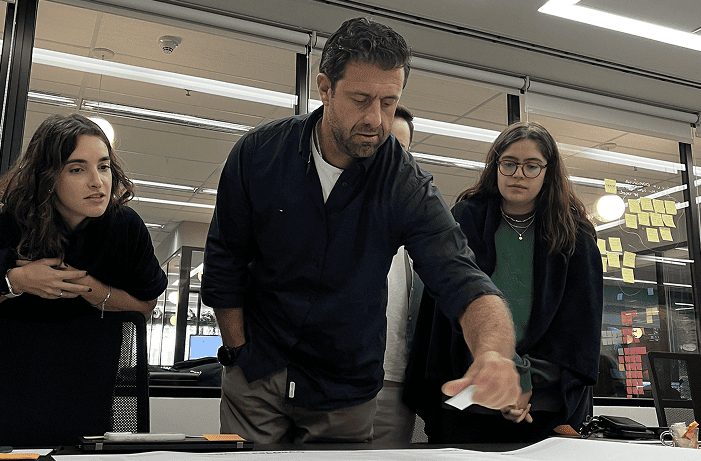 Homem em pé interagindo com materiais sobre uma mesa durante uma sessão de trabalho colaborativo, acompanhado por outras pessoas.