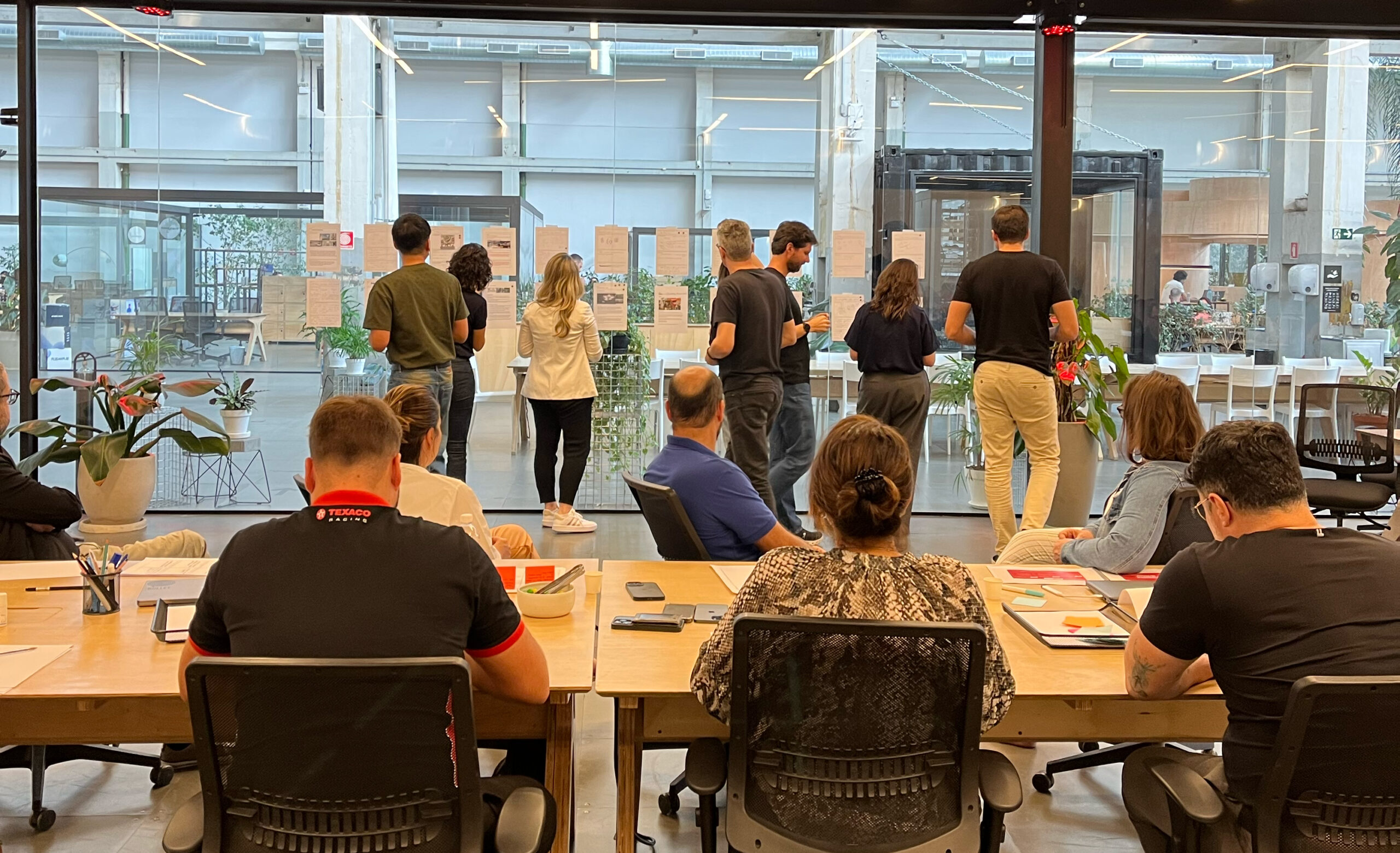 Participantes de um workshop analisam materiais expostos em uma parede de vidro, enquanto outras pessoas permanecem sentadas ao redor de mesas em um ambiente corporativo amplo e colaborativo.