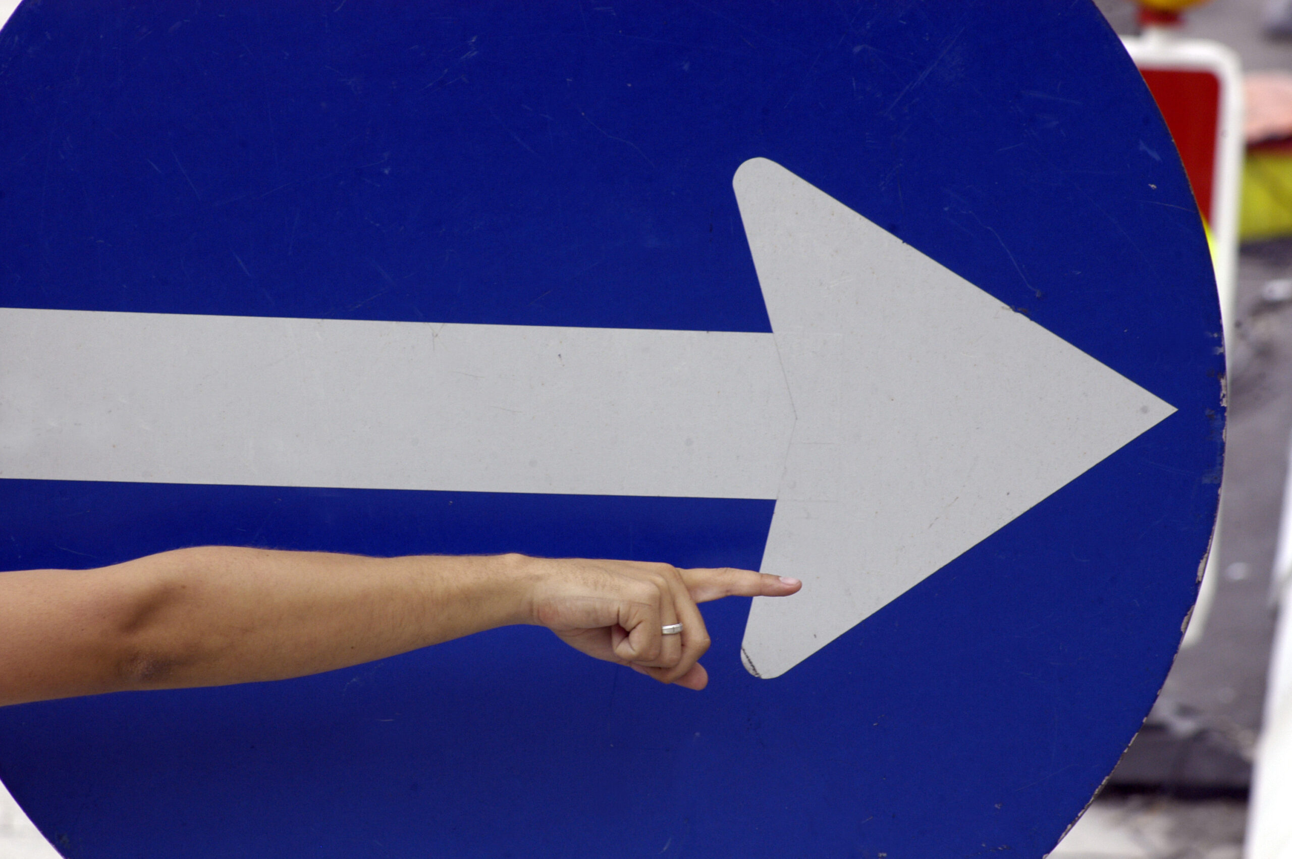Braço de uma pessoa aponta para a direita, acompanhando a direção indicada por uma grande seta branca sobre uma placa circular azul.