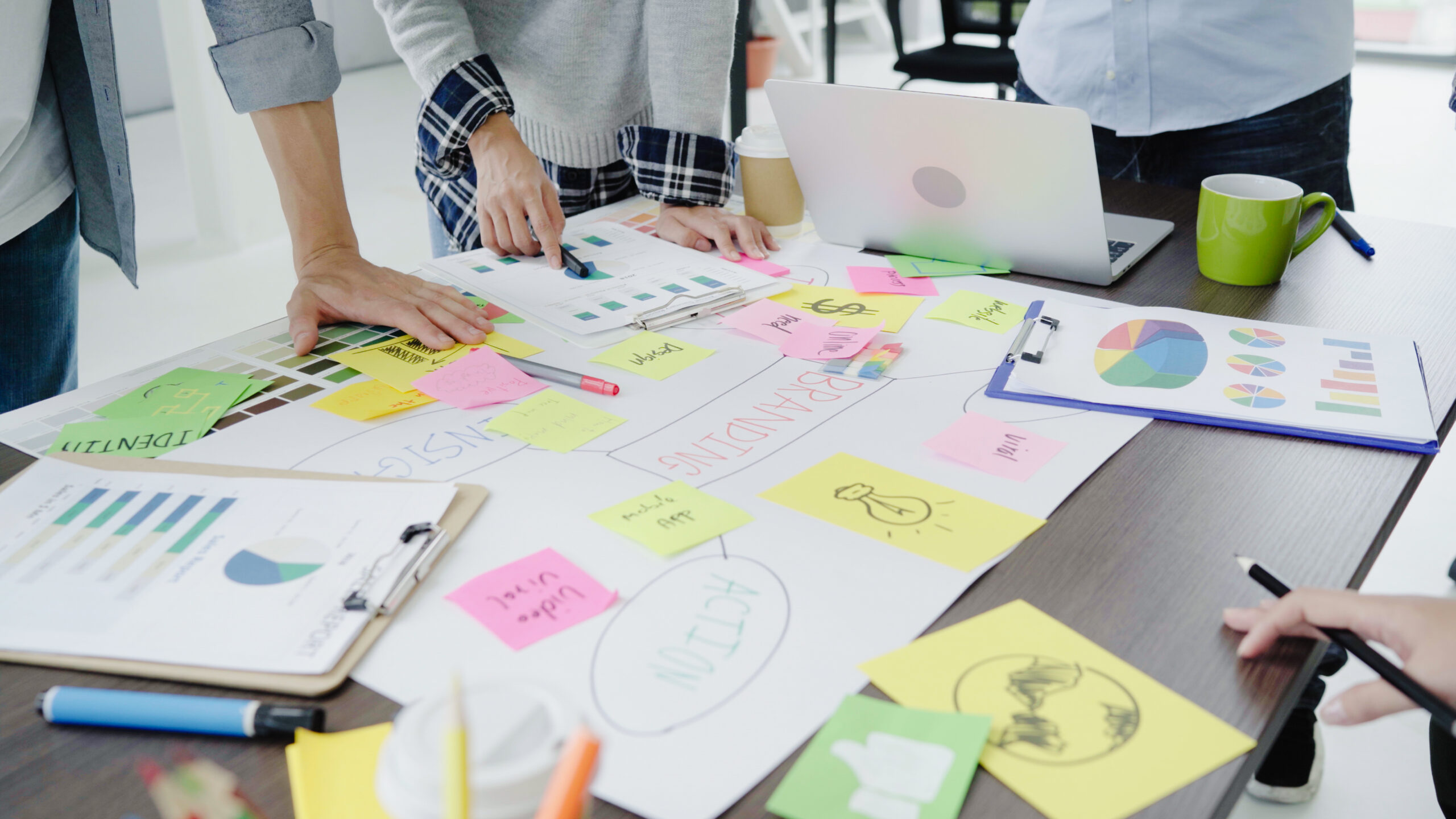 Pessoas colaboram em torno de uma mesa com post-its, gráficos, anotações e um notebook, durante uma atividade de planejamento e organização de ideias.