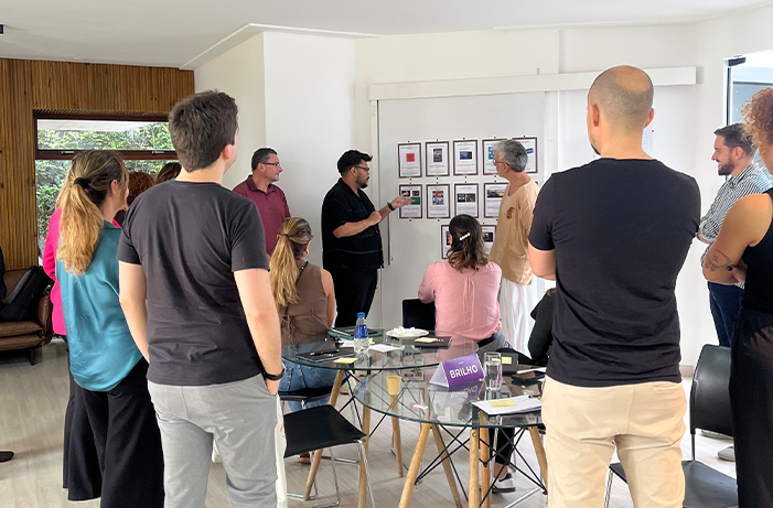 Grupo de pessoas acompanha a apresentação de ideias em um painel, durante uma atividade colaborativa em sala de reunião.