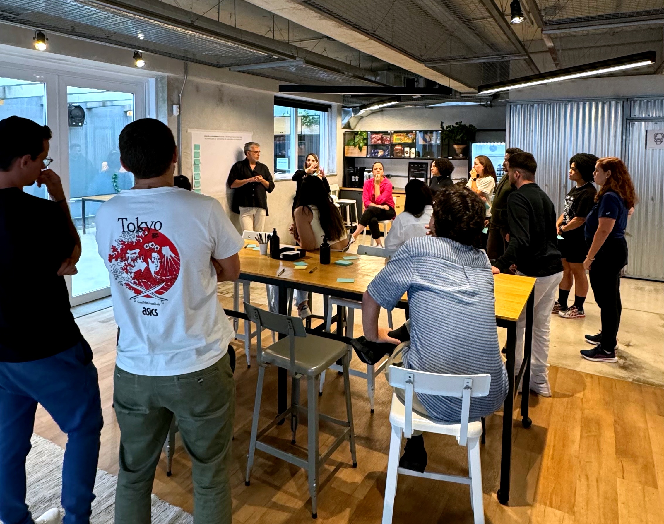 Fotografia de uma atividade colaborativa de endomarketing em ambiente corporativo. Um grupo de profissionais está reunido ao redor de mesas, ouvindo a apresentação de dois facilitadores. O espaço conta com materiais de apoio, quadro para anotações e post-its, reforçando uma dinâmica de escuta, troca de ideias e construção coletiva.