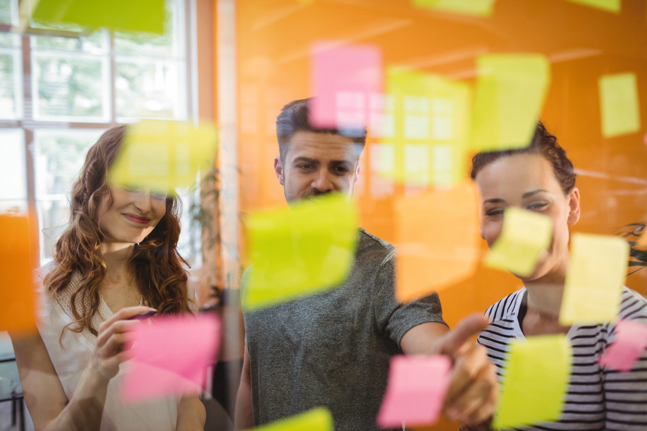 Fotografia de uma dinâmica colaborativa em ambiente corporativo. Três profissionais analisam e organizam post-its coloridos fixados em uma parede de vidro, representando troca de ideias, planejamento conjunto, priorização e construção coletiva de soluções.