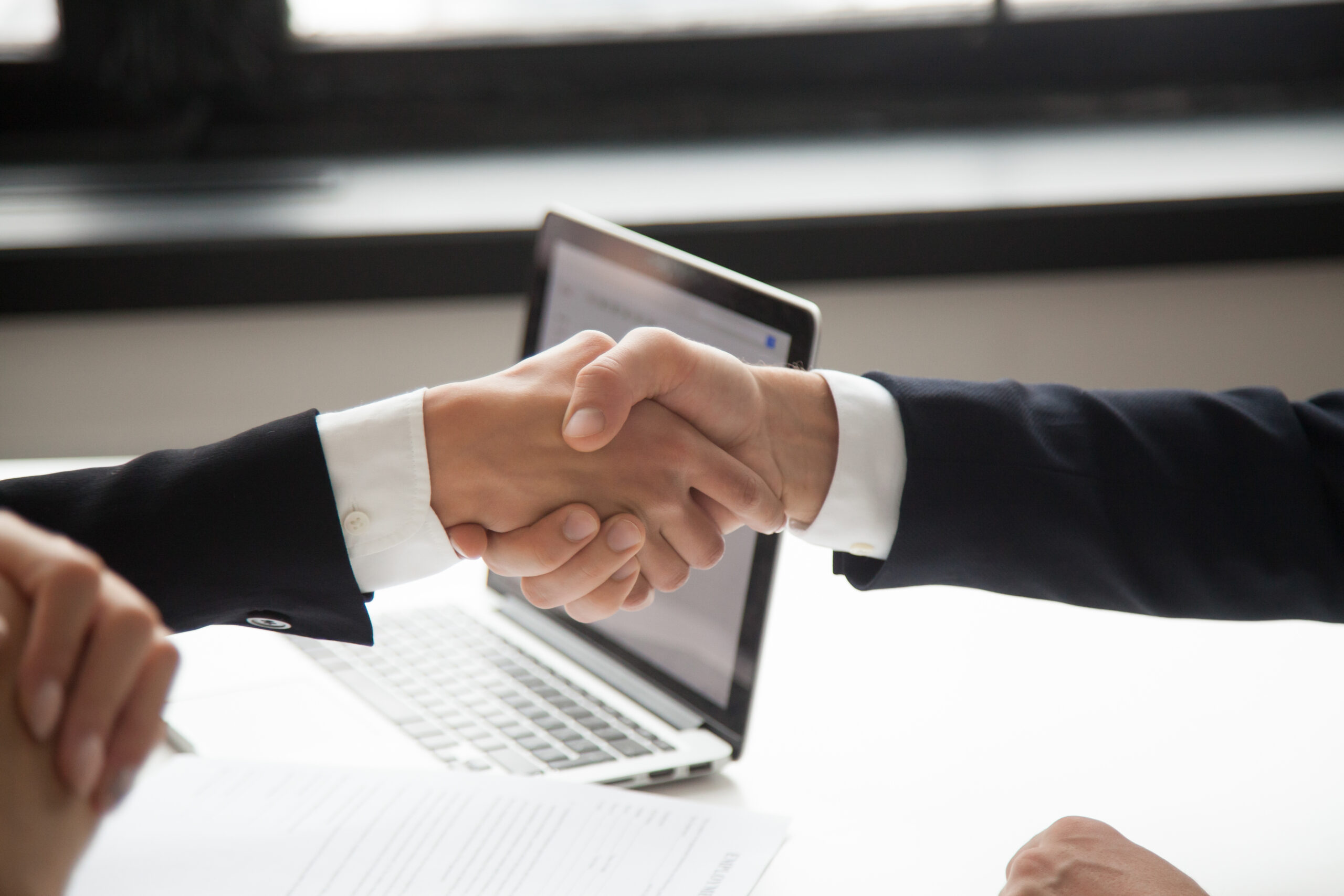 Duas pessoas de terno apertam as mãos sobre uma mesa de trabalho, com um notebook aberto ao fundo, simbolizando acordo, parceria ou negociação profissional.